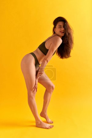 Photo for Full-length portrait of positive young woman fit slim body, slender legs, sitting in underwear against yellow studio background. Concept of female beauty, body care, fitness, sport, health, figure, ad - Royalty Free Image