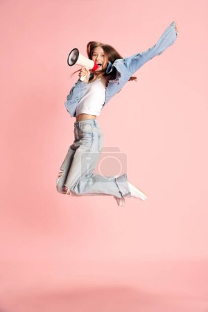 Téléchargez les photos : Portrait complet de jeune belle fille, étudiante en tenue décontractée sautant et criant en mégaphone sur fond de studio rose. Concept d'émotions humaines, mode, jeunesse, mode de vie, beauté, publicité - en image libre de droit