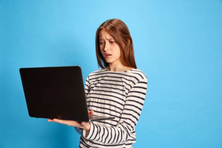 Photo for Error. Portrait of young girl in casual clothes looking with worried face in laptop over blue studio background. Concept of business, education, emotions, fashion, youth, lifestyle, female beauty, ad - Royalty Free Image