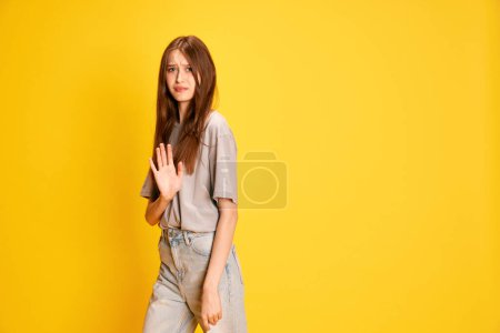 Téléchargez les photos : Rejet. Jeune fille, étudiante en tenue décontractée debout avec geste de déclin sur fond de studio jaune. Concept d'émotions humaines, mode, jeunesse, mode de vie, beauté féminine, publicité - en image libre de droit