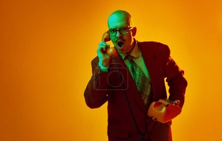 Photo for Portrait of young bald man with moustache in suit emotionally talking on retro phone against orange studio background in neon light. Concept of human emotions, facial expression, lifestyle - Royalty Free Image