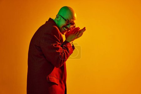 Photo for Portrait of young bald man with moustache emotionally rubbing hands against orange studio background in neon light. Concept of human emotions, facial expression, lifestyle - Royalty Free Image