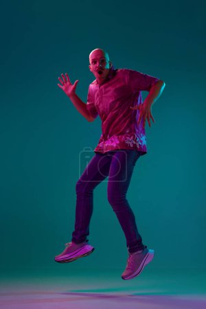 Photo for Full-length portrait of young bald man with moustache in stylish clothes emotionally jumping against cyan studio background in neon light. Concept of human emotions, facial expression, lifestyle - Royalty Free Image