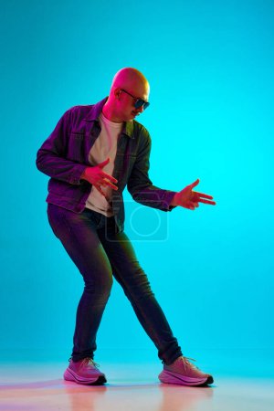 Photo for Full-length portrait of young bald man with moustache wearing casual clothes, posing against blue studio background in neon light. Concept of human emotions, facial expression, lifestyle - Royalty Free Image