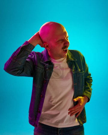 Photo for Portrait of young bald man with moustache standing with thoughtful face against blue studio background in neon light. Headache. Concept of human emotions, facial expression, lifestyle - Royalty Free Image