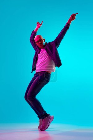 Photo for Full-length portrait of young bald man with moustache in casual clothes dancing against blue studio background in neon light. Concept of human emotions, facial expression, lifestyle - Royalty Free Image