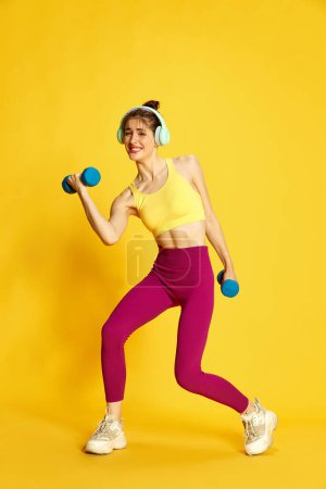 Photo for Full-length image of young slim woman in sportswear and headphones training with heavy dumbbells against yellow studio background. Concept of sport, fitness, body care, fashion, youth, lifestyle, ad - Royalty Free Image