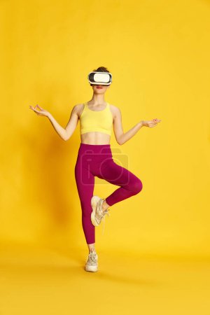 Photo for Full-length image of young slim woman wearing vr glases and training against yellow studio background. Yoga and meditation. Concept of sport, fitness, body care, fashion, youth, lifestyle, ad - Royalty Free Image