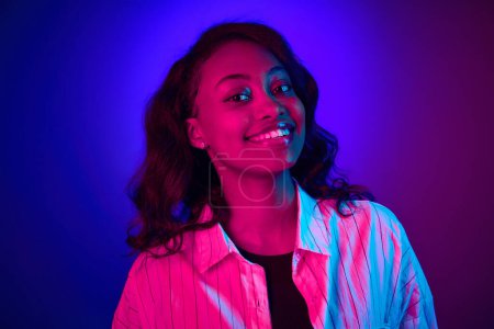 Photo for Close-up portrait of beautiful, young, african woman looking at camera, smiling against gradient blue purple studio background in neon light. Concept of human emotions, youth, fashion, lifestyle, ad - Royalty Free Image