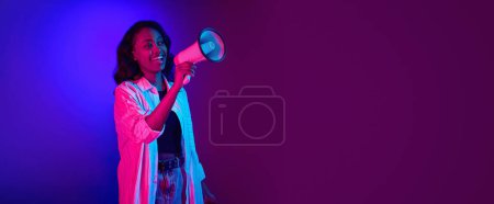 Portrait of young, smiling, african woman talking in megaphone against gradient blue purple studio background in neon light. News. Concept of human emotions, youth, fashion, lifestyle, ad. Banner