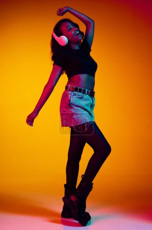 Photo for Full-length portrait of beautiful, young, african, stylish woman listening to music in headphones and dancing over yellow background in neon light. Concept of emotions, youth, fashion, lifestyle, ad - Royalty Free Image