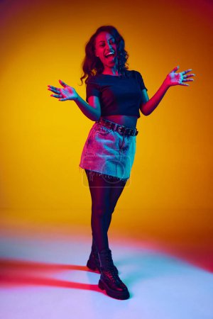 Photo for Full-length portrait of young beautiful african woman in stylish clothes cheerfully clapping hands against yellow studio background in neon light. Concept of human emotions, youth, fashion, lifestyle - Royalty Free Image