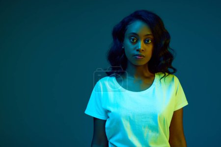 Photo for Portrait of young, african, beautiful woman in white t-shirt posing, looking at camera with serious face against cyan, blue background in neon light. Concept of emotions, youth, fashion, lifestyle, ad - Royalty Free Image