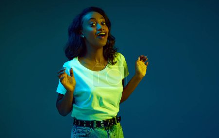 Photo for Portrait of young, african, beautiful woman in white t-shirt posing with excited face against cyan, blue studio background in neon light. Concept of human emotions, youth, fashion, lifestyle, ad - Royalty Free Image