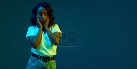 Foto de Retrato de una hermosa, joven, mujer africana en camiseta blanca y falda de jeans posando contra cian, fondo estudio azul en luz de neón. Concepto de emociones humanas, juventud, moda, estilo de vida, anuncio - Imagen libre de derechos