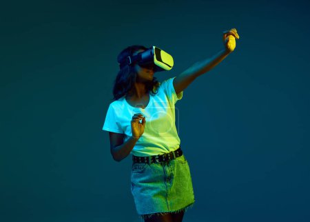 Photo for Portrait of attractive young, african woman in white t-shirt wearing vr glasses, posing against cyan, blue studio background in neon light. Concept of human emotions, youth, fashion, lifestyle, ad - Royalty Free Image