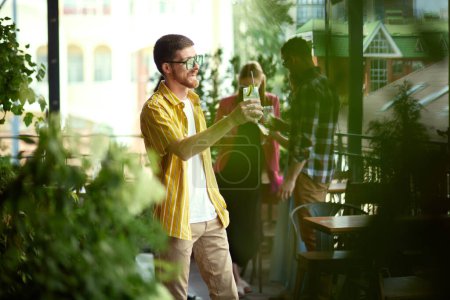 Photo for Focus on cheerful man in stylish colorful clothes and sunglasses, raising cocktail, meeting with friends in cafe. Blurred people on background. Friendship, party, communication, alcohol drinks concept - Royalty Free Image