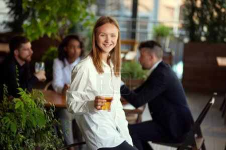 Téléchargez les photos : Concentrez-vous sur une jeune employée souriante debout avec de la bière. Des gens flous sur le fond. Rencontre avec l'équipe. Concept de temps libre, d'affaires, de communication, d'amusement, de boisson alcoolisée - en image libre de droit