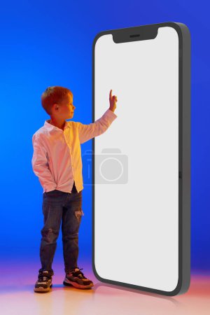 Foto de Imagen completa de niño pequeño, niño desplazándose en 3D pantalla del teléfono móvil contra fondo azul estudio en luz de neón. Concepto de infancia, estilo de vida, emociones, educación, internet, anuncio - Imagen libre de derechos