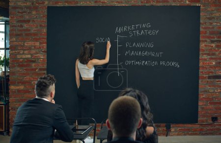 Photo for Young woman, business tutor writing on blackboard about marketing tools. Meeting with trainees, workers. Business education. Concept of business, planning, strategy, brainstorming, analytics, ad - Royalty Free Image