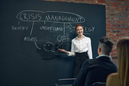 Photo for Young woman, employee writing on black board plan of crisis management. Meeting with colleagues for discussion and planning. Concept of business, planning, strategy, brainstorming, analytics, ad - Royalty Free Image