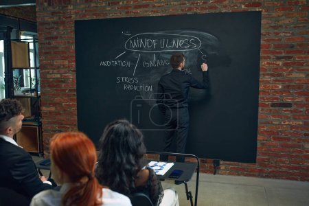Photo for Young man writing on blackboard tips and components of mindfulness set up. Meeting with workers, colleagues. Psychology. Concept of business, planning, strategy, brainstorming, analytics, ad - Royalty Free Image