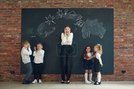 Photo for Young cheerful woman in formal wear standing by blackboard with drawn icons and playing with children. Education and game. Concept of business, education, lessons, games, study, psychology, ad - Royalty Free Image