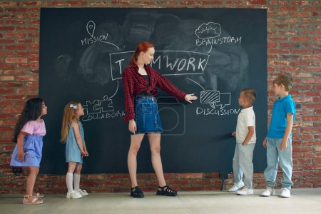 Photo for Young woman in casual clothes teaching kids to work together. Teamwork strategy icons on blackboard. Concept of business, education, lessons, games, study, psychology, ad - Royalty Free Image