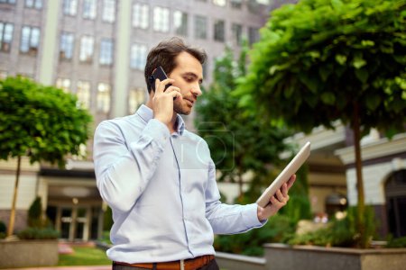 Photo for Young businessman standing outside office outdoors, looking on tablet and talking on mobile phone. Conversation. Concept of business, career development, ambitions, success, office lifestyle - Royalty Free Image