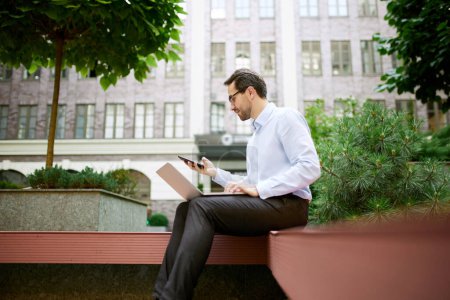 Photo for Young businessman sitting on bench outside of his office and working online with laptop and mobile phone. Concept of business, career development, ambitions, success, office lifestyle - Royalty Free Image