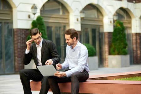 Photo for Two businessmen, coworkers sitting on bench outside the office, looking on laptop and working. Analyitcs. Concept of business, career development, ambitions, success, office lifestyle - Royalty Free Image