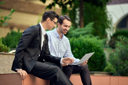 Photo for Two businessmen, coworkers sitting on bench outside the office, looking on laptop and working. Concept of business, career development, ambitions, success, office lifestyle - Royalty Free Image