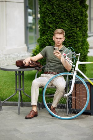Photo for Stylish, handsome, serious businessman sitting on bench and reading news, mails on mobile phone. Going to work on bike. Concept of business, active lifestyle, fashion, youth, ecology - Royalty Free Image