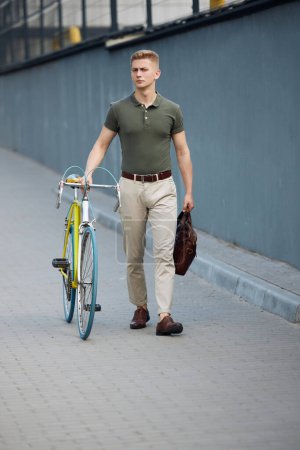 Foto de Imagen completa de un hombre de negocios joven y elegante, empleado con ropa casual inteligente caminando por la calle con bicicleta. Camino a la oficina. Negocios, estilo de vida activo, moda, juventud, concepto de ecología - Imagen libre de derechos