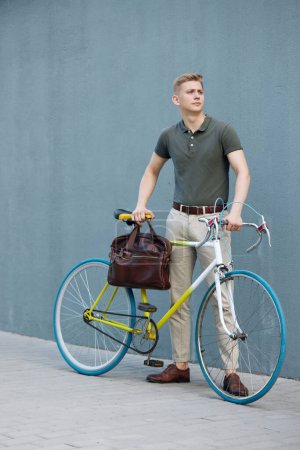 Foto de Guapo, concentrado, hombre de negocios con estilo en ropa casual inteligente y maletín de pie al aire libre con bicicleta. Voy a la oficina. Concepto de negocio, estilo de vida activo, moda, juventud, ecología - Imagen libre de derechos