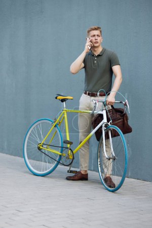 Photo for Handsome, concentrated, stylish businessman in smart casual clothes standing outdoors with bike and talking on mobile phone. Concept of business, active lifestyle, fashion, youth, ecology - Royalty Free Image