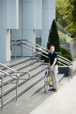 Photo for Handsome, young, stylish employee, worker in smart casual clothes standing near office building with bicycle. Going to work on bike. Concept of business, active lifestyle, fashion, youth, ecology - Royalty Free Image