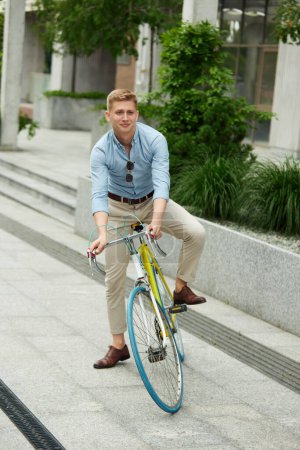 Foto de Joven alegre, positivo, guapo, empleado con ropa elegante que va a la oficina en bicicleta. Fondo urbano. Concepto de negocio, estilo de vida activo, moda, juventud, ecología - Imagen libre de derechos