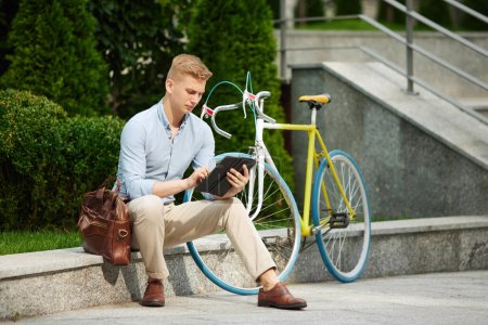 Foto de Joven empresario concentrado y guapo sentado en el banco fuera de la oficina con la bicicleta y trabajando en línea en la tableta. Concepto de negocio, estilo de vida activo, moda, juventud, ecología - Imagen libre de derechos
