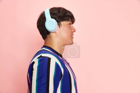 Téléchargez les photos : Profil portrait de jeune homme écoutant de la musique dans un casque et souriant sur fond de studio rose sur fond de studio rose. Concept d'émotions humaines, style de vie, mode décontractée, publicité - en image libre de droit