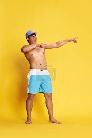 Photo for Full-length portrait of joyful, cheerful young man in swimming trunks, sunglasses and cap posing over yellow studio background. Concept of summer, vacation, leisure time, holidays, human emotions, ad - Royalty Free Image