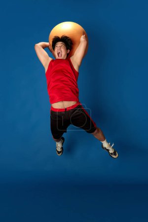 Photo for Full-length portrait of young ma in sportswear training, jumping with fitness ball against blue studio background. Concept of sport, fitness, active and healthy lifestyle, human emotions, ad - Royalty Free Image