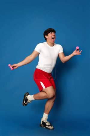Photo for Full-length portrait of young man in sportswear training with dumbbells and having fun against blue studio background. Concept of sport, fitness, active and healthy lifestyle, human emotions, ad - Royalty Free Image