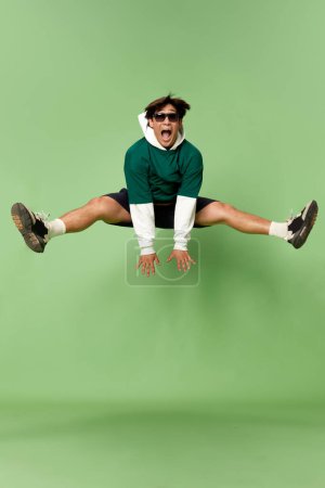 Téléchargez les photos : Portrait de jeune homme élégant en lunettes de soleil et vêtements de sport sautant joyeusement sur fond de studio vert. Concept d'émotions humaines, style de vie, années 90, mode, joie, jeunesse, publicité - en image libre de droit