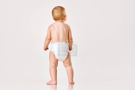 Photo for Back view image of little baby, child, toddler standing in diaper against white studio background. Concept of childhood, newborn lifestyle, happiness, care. Copy space for ad - Royalty Free Image