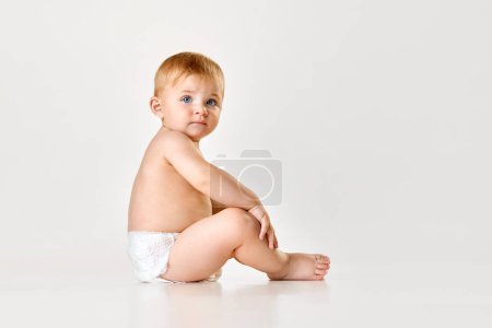Foto de Hermoso bebé, niño pequeño en pañal sentado en el suelo contra el fondo blanco del estudio. Calma, niña. Concepto de infancia, estilo de vida del recién nacido, felicidad, cuidado. Copiar espacio para anuncio - Imagen libre de derechos