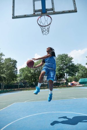 Téléchargez les photos : Image pleine longueur d'un jeune homme compétitif en uniforme bleu jouant au basket, lançant une balle. Terrain de sport extérieur. Concept de sport professionnel, compétition, passe-temps, jeu, mode de vie actif - en image libre de droit