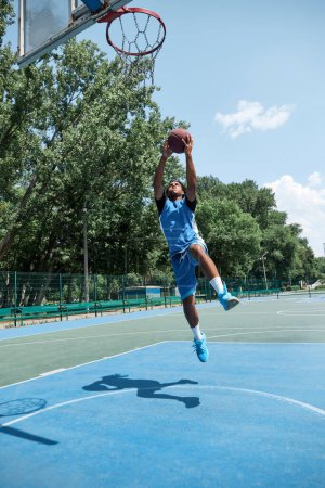 Photo for Full-length dynamic image of young guy in blue uniform jumping, playing basketball outdoors on sportsground on warm sunny day. Concept of professional sport, competition, hobby, game, active lifestyle - Royalty Free Image