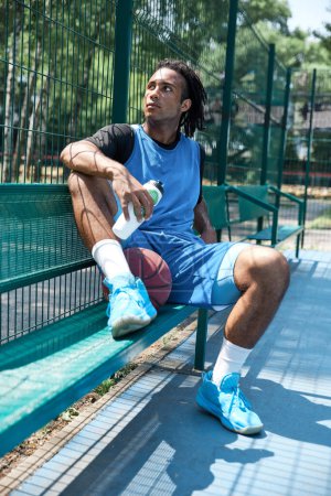 Téléchargez les photos : Jeune homme avec des dreads en uniforme bleu assis sur le banc après avoir joué au basket-ball sur terrain de sport extérieur. Je me repose. Concept de sport professionnel, compétition, passe-temps, jeu, mode de vie actif - en image libre de droit