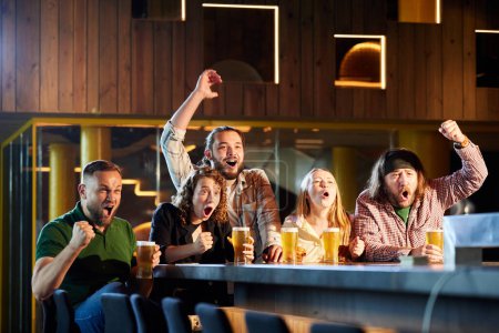 Foto de Amigos, hombres y mujeres que asisten a pub, bar, viendo traducción de partidos deportivos, bebiendo cerveza, animando a su equipo favorito. Concepto de competición deportiva, hobby, estilo de vida, emociones humanas, diversión - Imagen libre de derechos